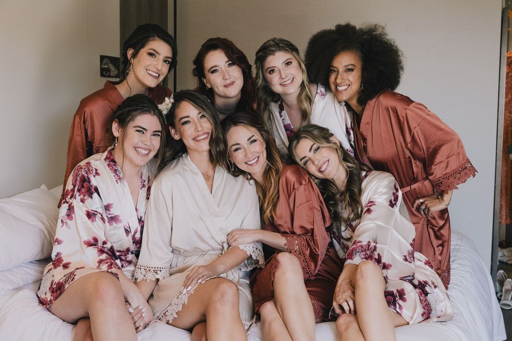 Bride and seven bridesmaids in satin robes smiling together on a white bed before the wedding.