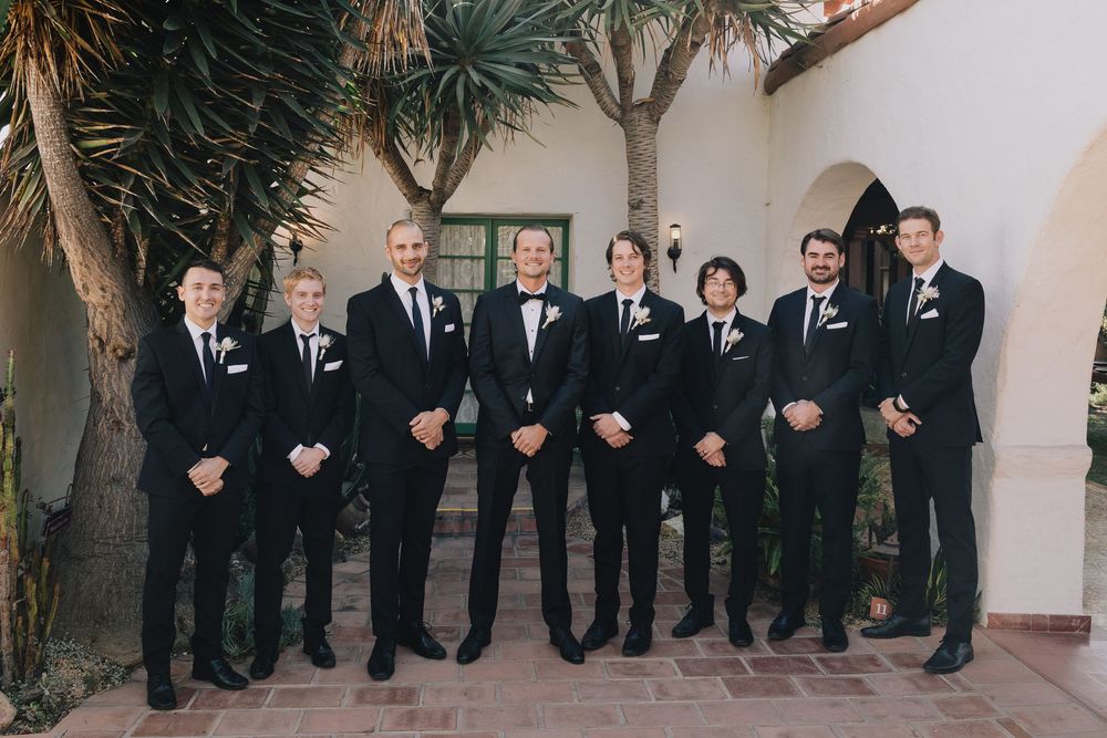 Eight groomsmen in matching black suits with boutonnieres pose together outdoors at a wedding venue.