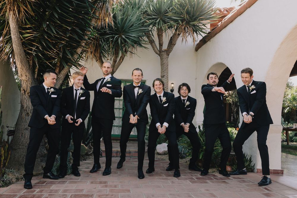 Groom and groomsmen in matching black tuxedos posing playfully in a courtyard with palm trees.