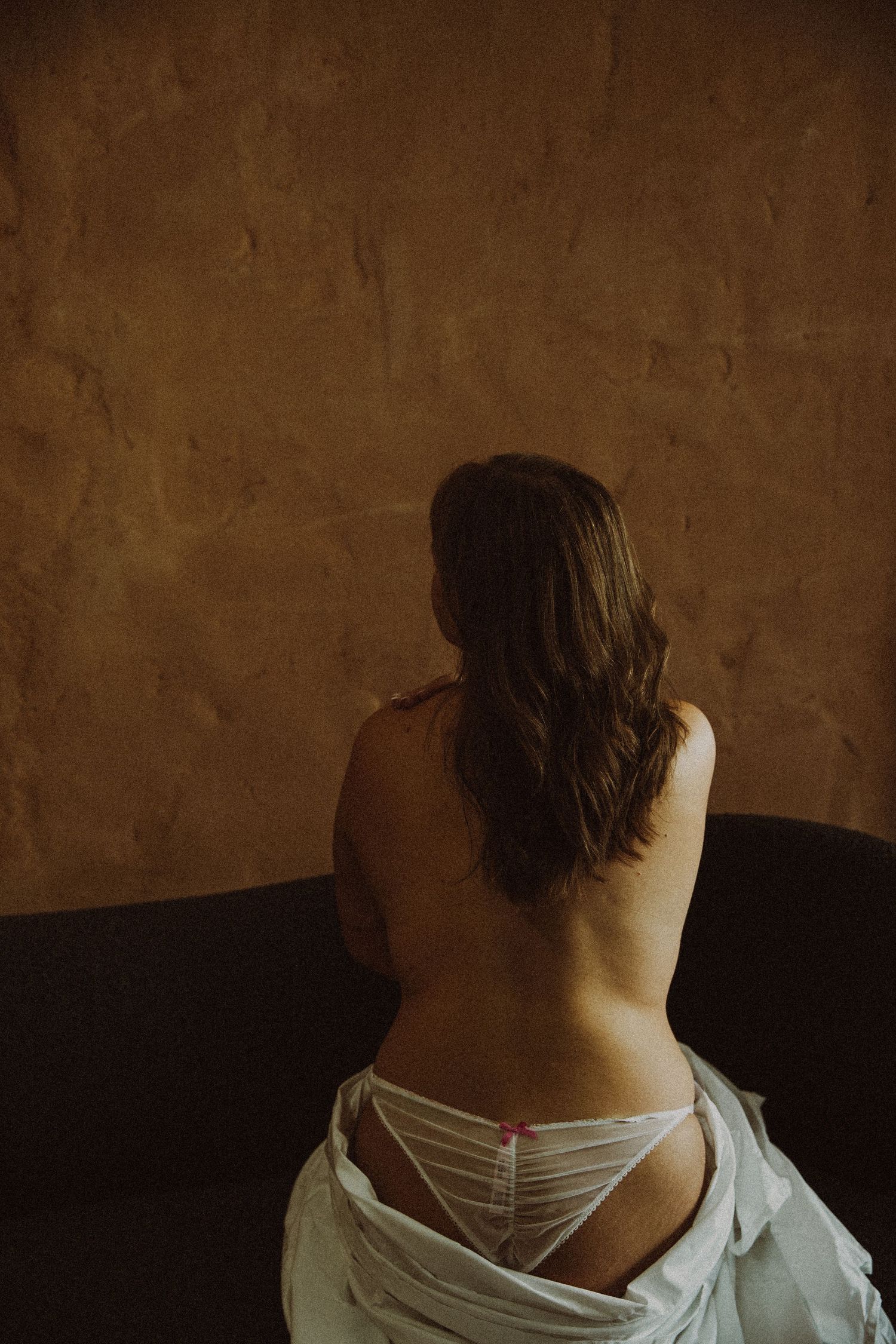 Woman with long brown hair seen from behind, sitting on dark sofa wearing white lingerie against textured brown wall.