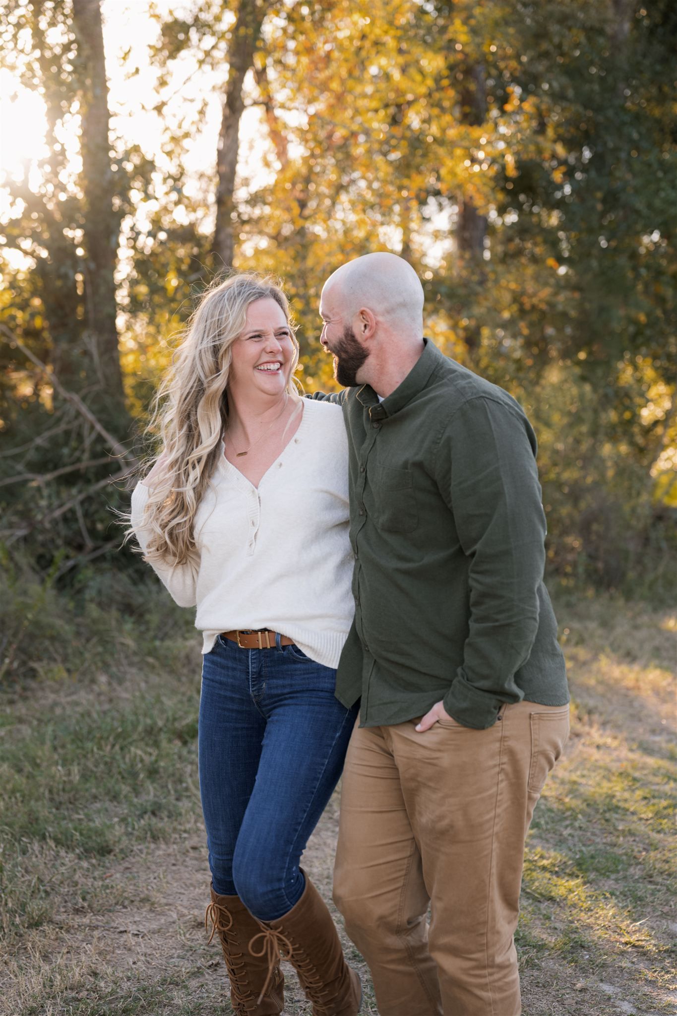 A smiling couple shares a warm moment outdoors at golden hour, surrounded by autumn trees on a dirt path.