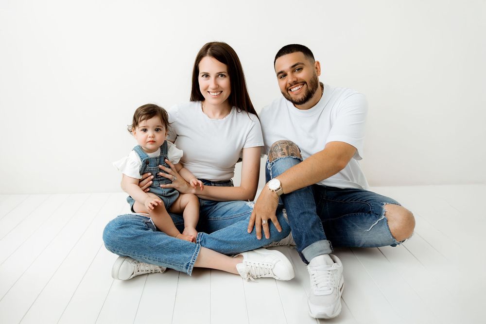 Family sitting on a studio flor wearing jeans and white t-shirts. Baby is on mom's lap