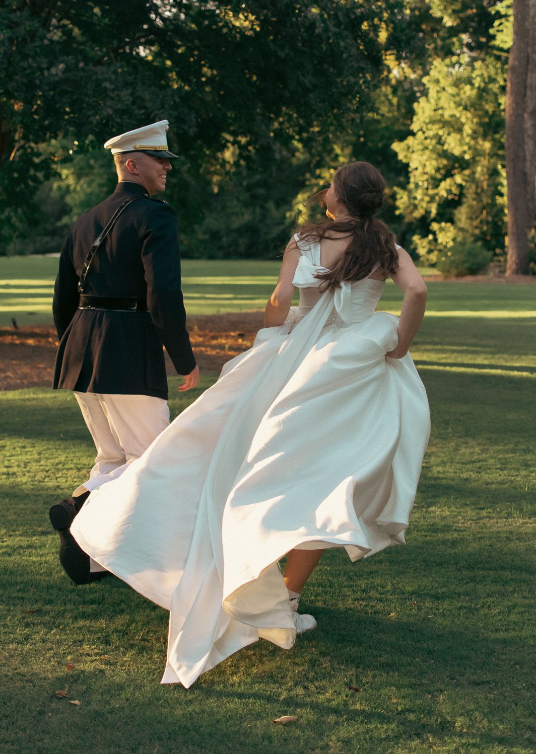 A couple in wedding attire dances playfully in a sunlit park during golden hour, with the bride's white dress flowing.