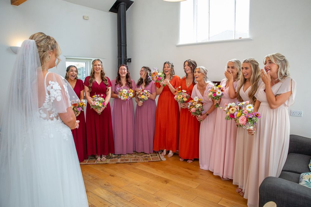 Bride reveals wedding dress to emotional bridesmaids in colorful red and pink gowns holding floral bouquets.