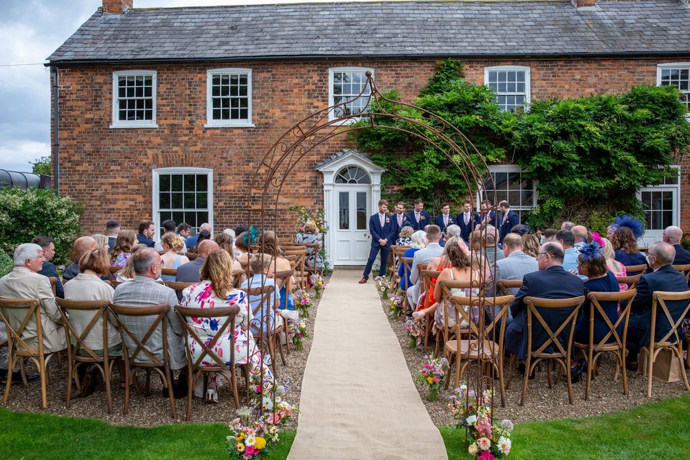 Outdoor wedding ceremony in front of brick farmhouse with guests seated along flower-lined aisle.