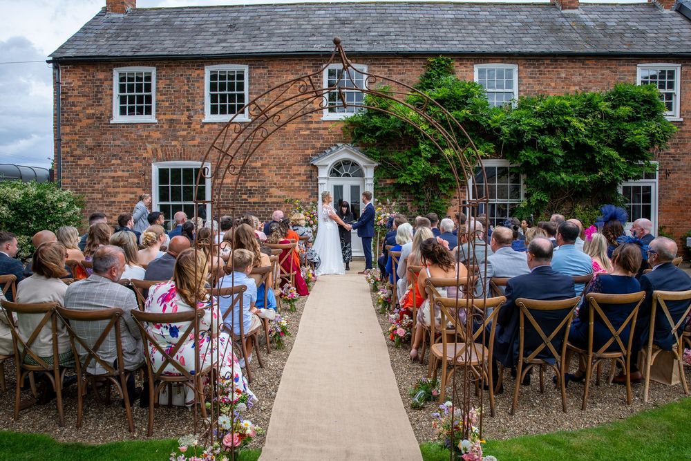 Bride and groom exchange vows at outdoor ceremony beneath floral arch in front of charming brick manor.
