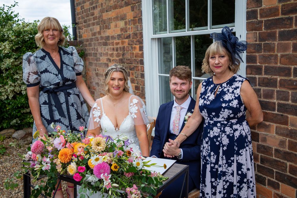 Bride and groom sign wedding register with two female witnesses beside colorful bridal bouquet outdoors.