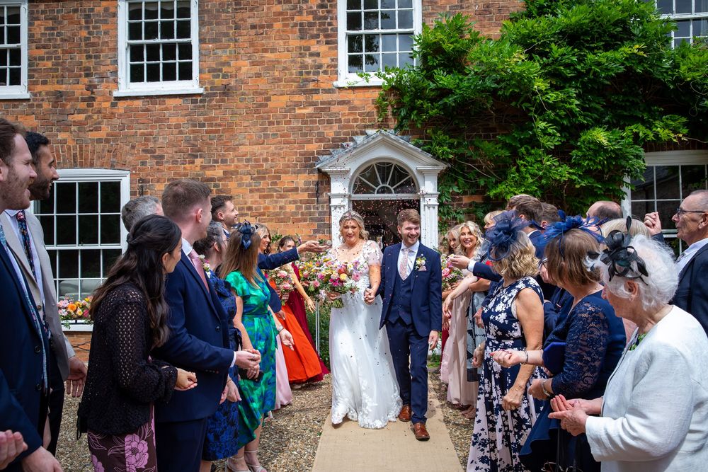 Bride and groom walk through confetti shower outside historic brick venue surrounded by cheering wedding guests.