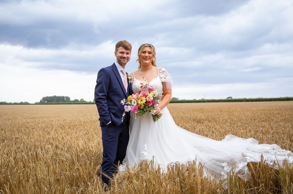 Smiling bride and groom pose together in golden wheat field, bride holding vibrant colorful bouquet.