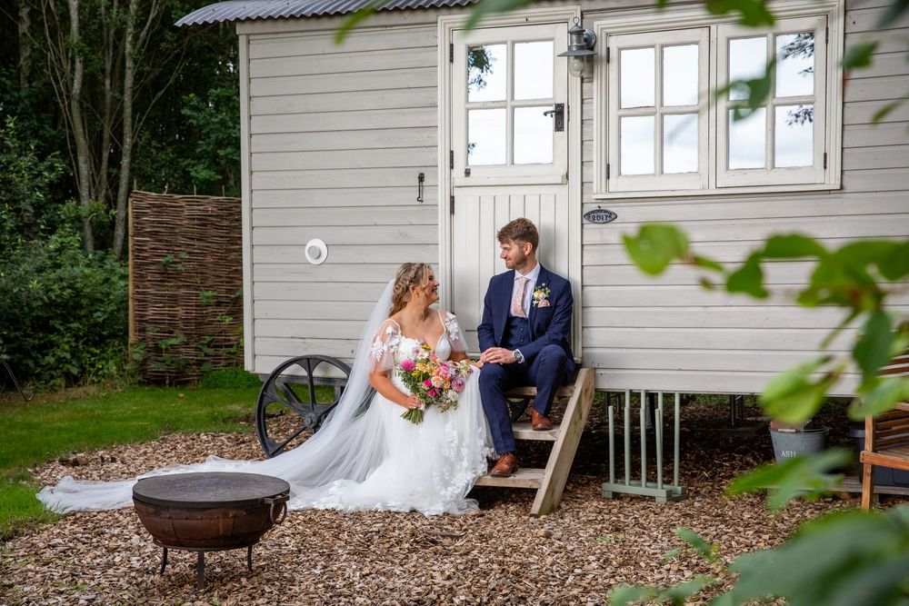 Bride and groom sit together on steps of charming white shepherd hut surrounded by lush garden.
