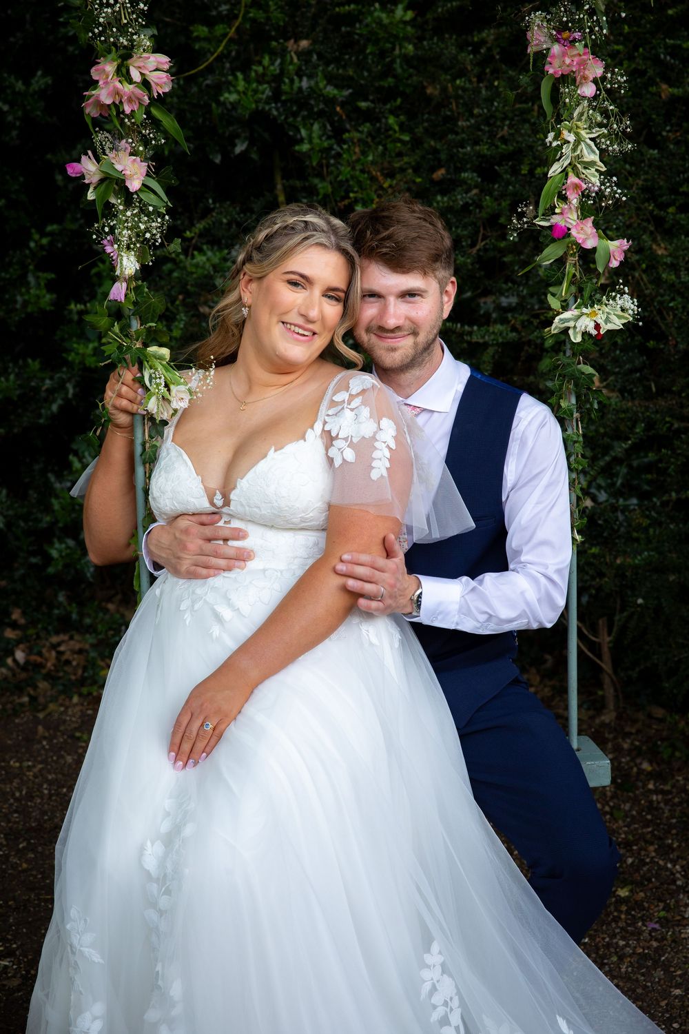 Bride and groom pose on floral decorated garden swing, bride in lace ballgown holding pink drink.