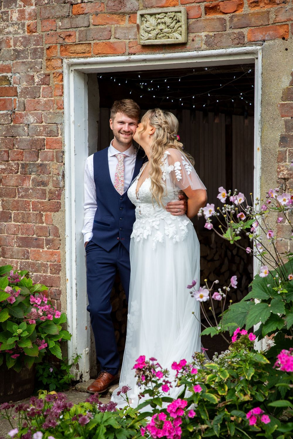 Bride and groom share romantic moment in rustic brick doorway surrounded by colorful garden flowers.