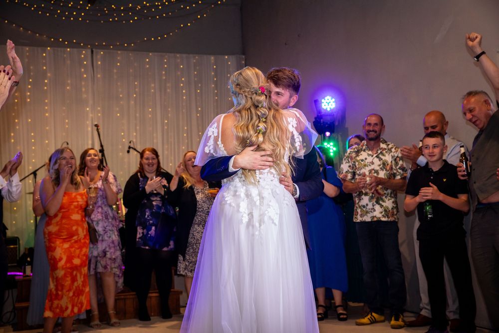 Bride and groom share first dance embracing on dance floor with cheering guests behind them.