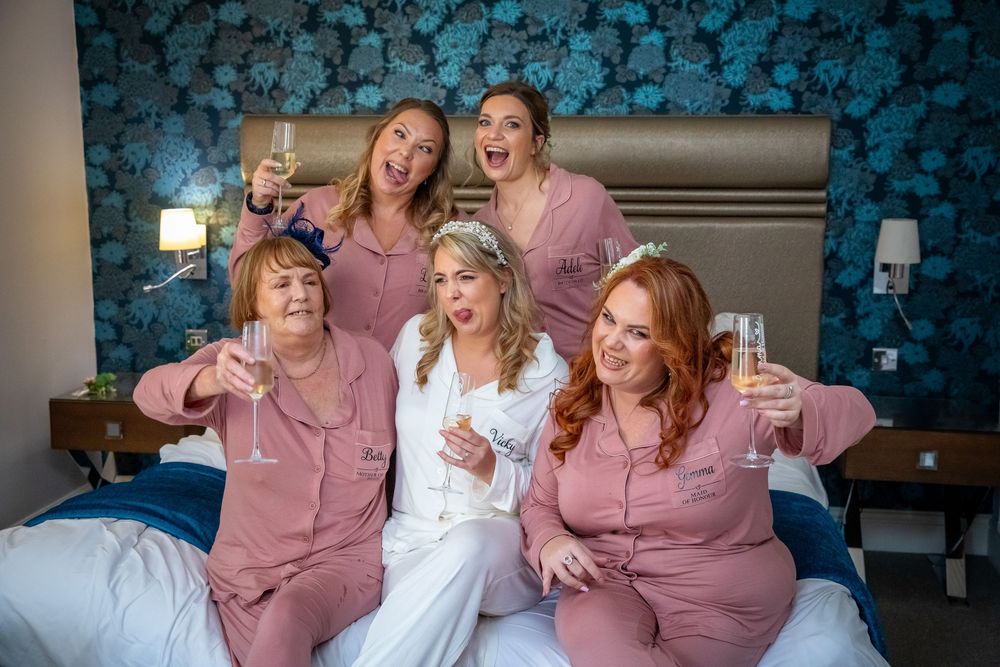 Bride and bridesmaids in pink pajamas celebrating with champagne on a bed before the wedding.