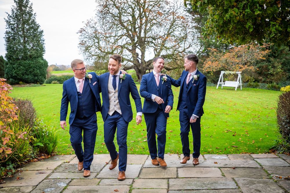 Four groomsmen in navy suits walking together outdoors in an autumn garden setting.