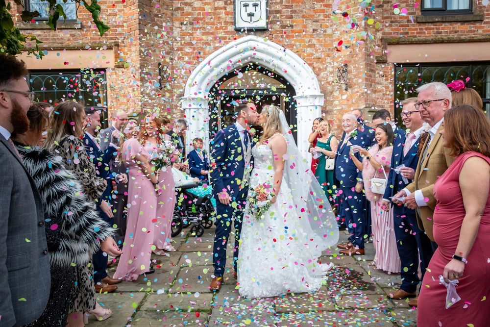 Bride and groom kissing in a shower of colorful confetti outside a brick wedding venue.