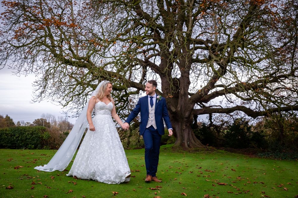 Bride and groom holding hands while walking across a lawn beside a large bare tree.