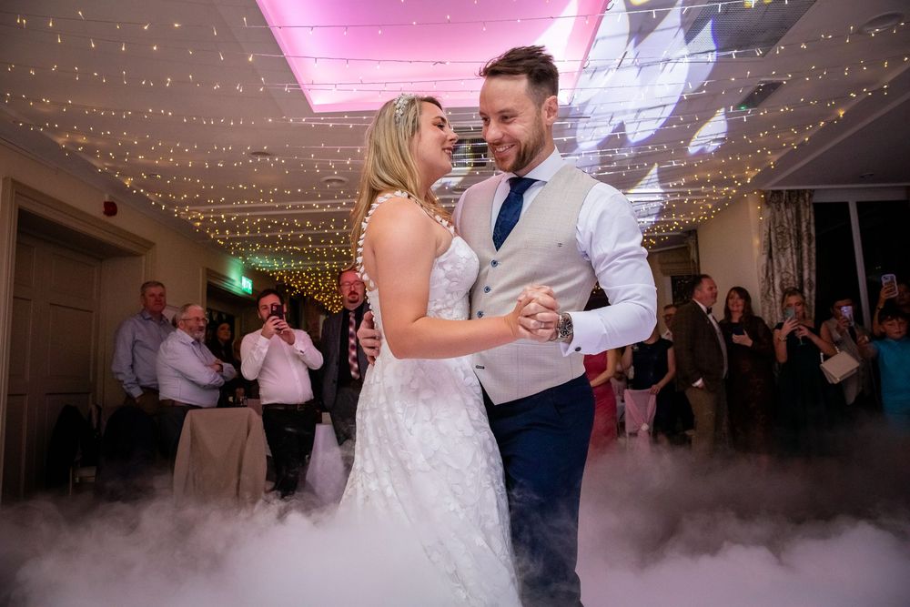 Bride and groom share their first dance surrounded by fairy lights and dry ice fog at elegant wedding reception.