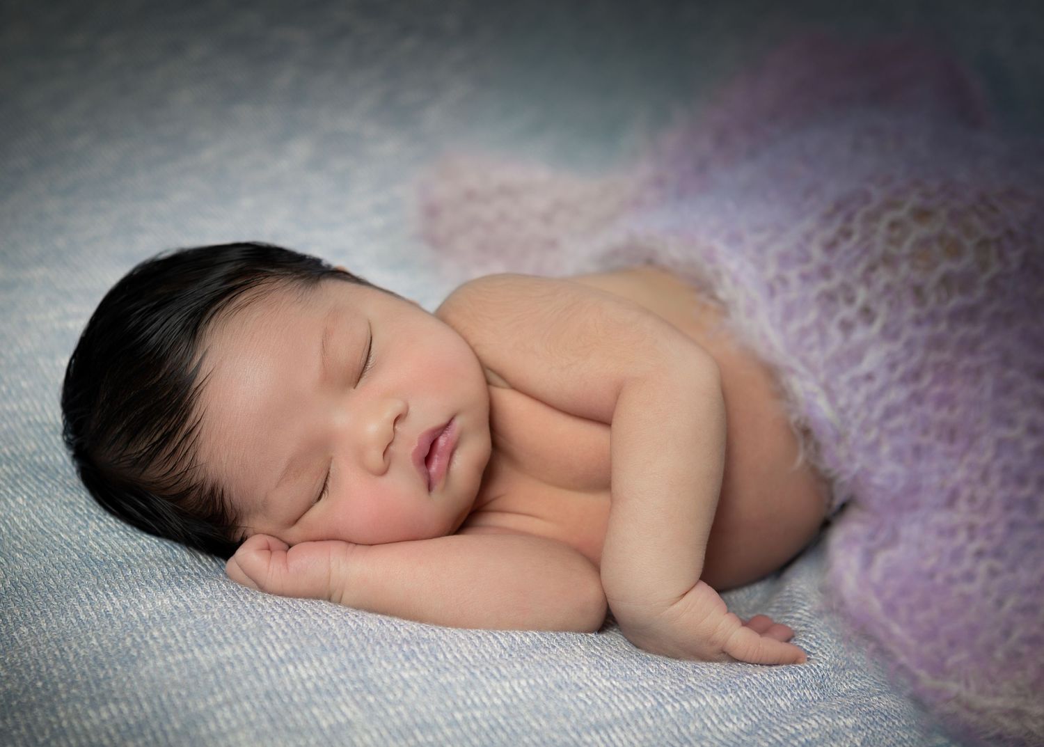 A sleeping newborn baby lies peacefully on soft pink and purple textured fabric in a professional photo series.