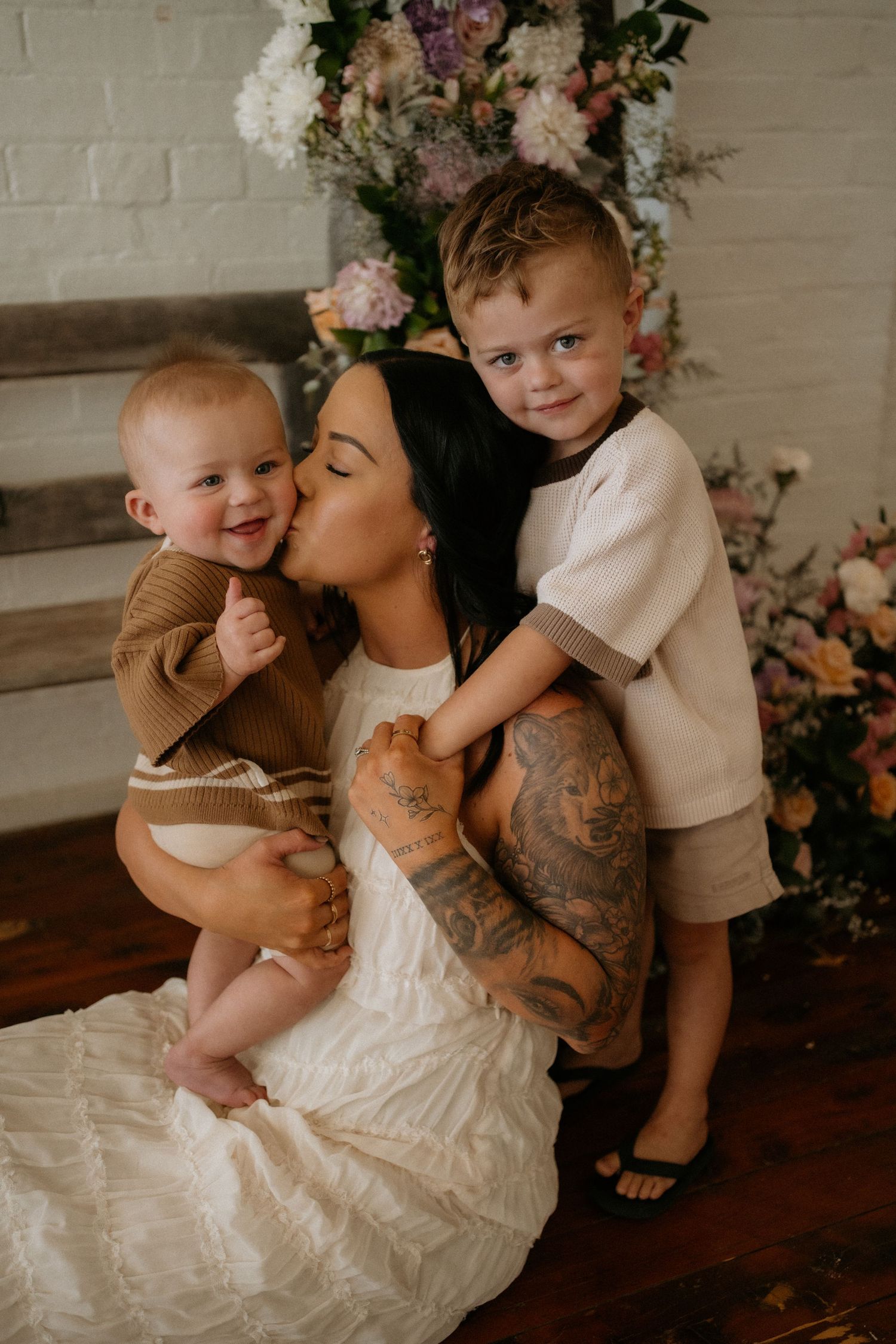 A tattooed mother in a white dress kisses her toddler son while holding a smiling baby, posed before a floral backdrop.