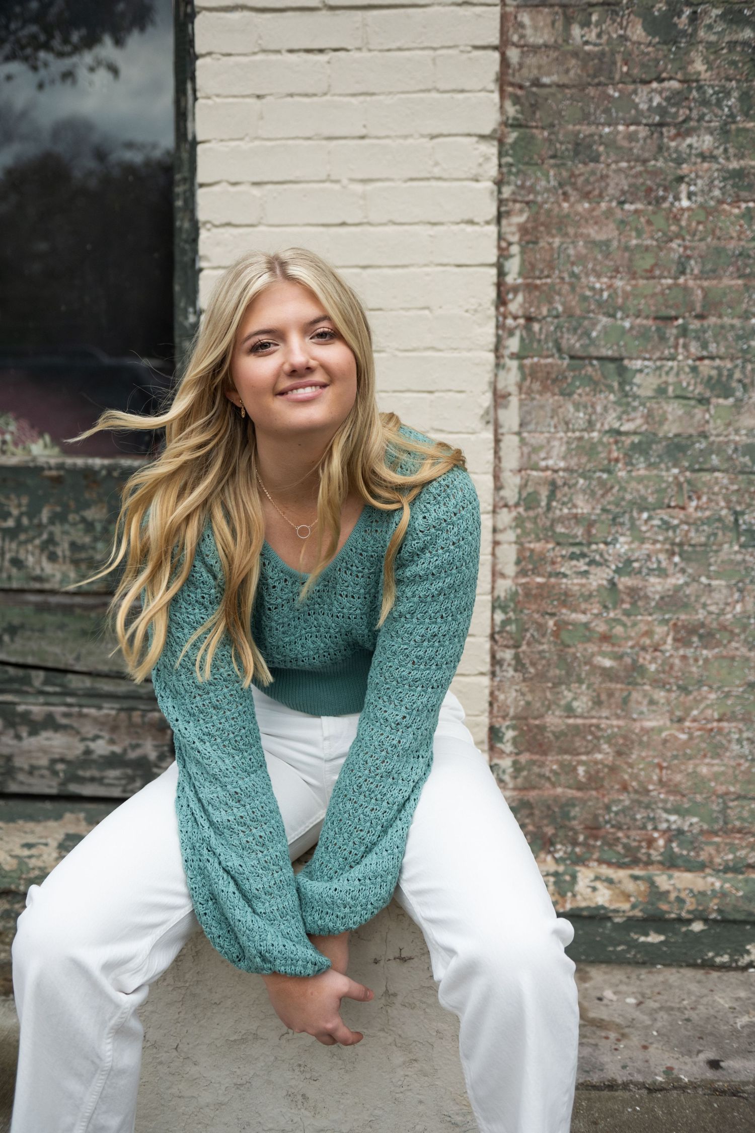 Person wearing a teal knit sweater and white pants poses playfully against a rustic brick wall.
