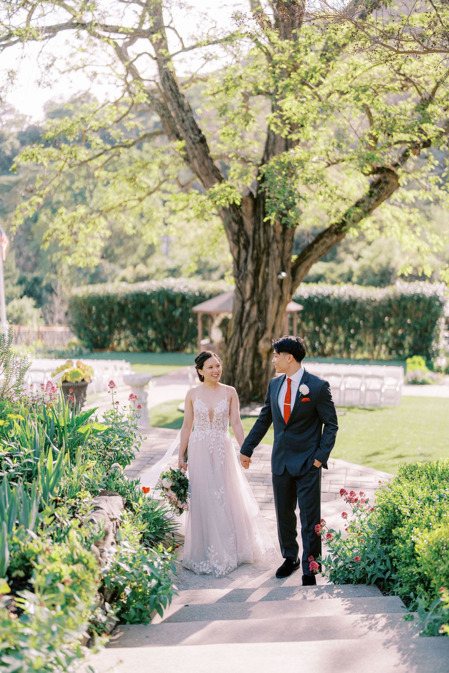 A couple walks together through a lush garden path lined with blooming flowers and trees on their wedding day.