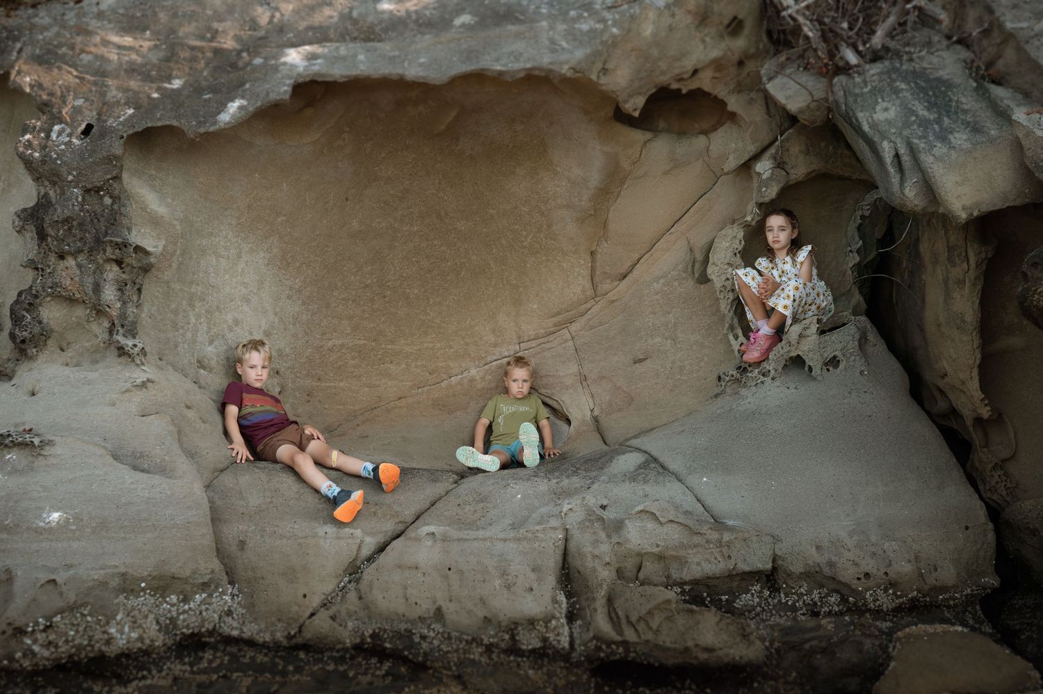 Three children sitting and playing on large smooth rocks inside a natural cave formation.
