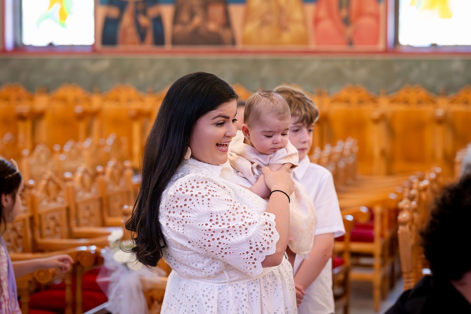 Woman in white lace dress holds baby in church during baptism ceremony with wooden pews in background.