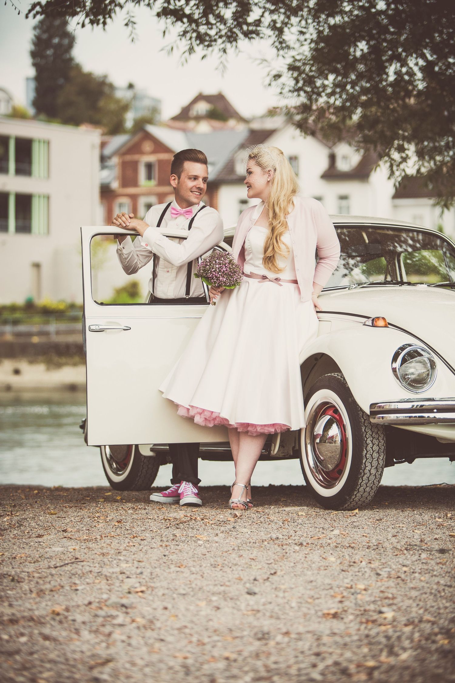 Couple in vintage style clothing posing beside a classic white VW Beetle car outdoors.