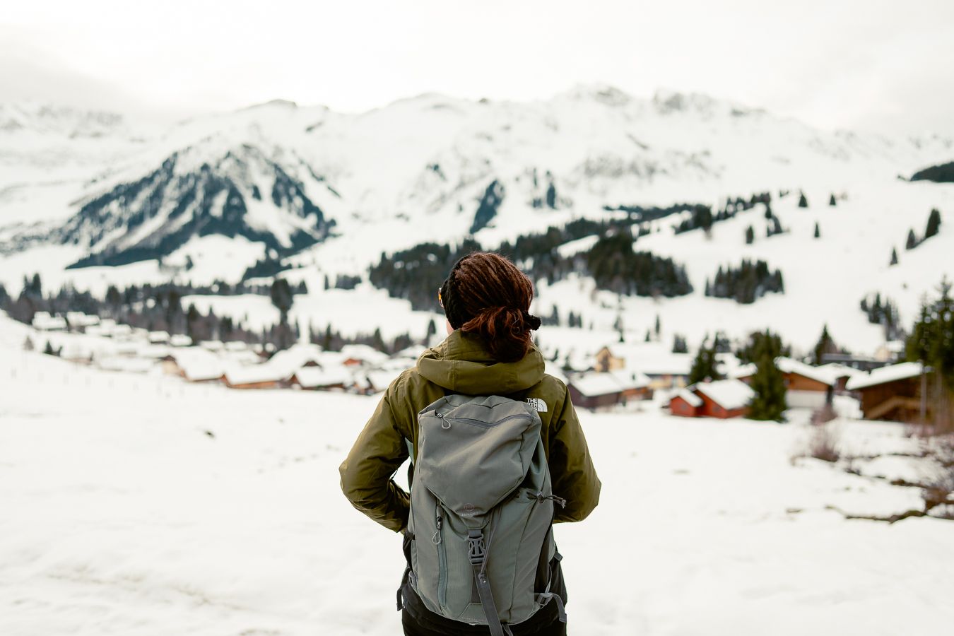 Hikerin mit Rucksack blickt auf das verschneite Dorf Sörenberg und die Gipfel der Schweizer Alpen unter bewölktem Himmel.