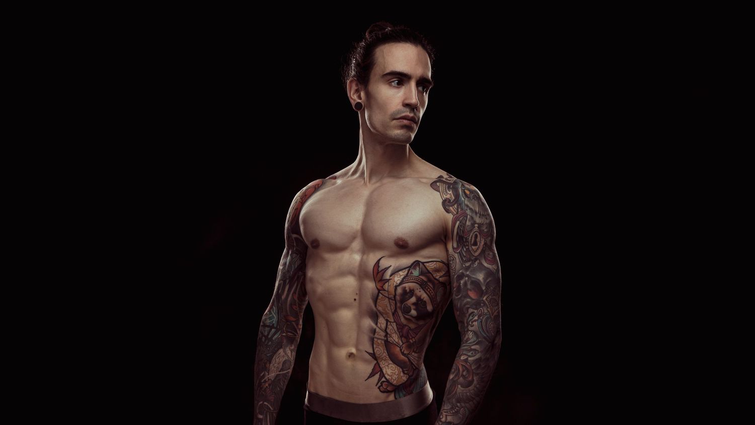 Tattooed shirtless muscular man posing against dark background showcasing extensive body art on arms and torso.