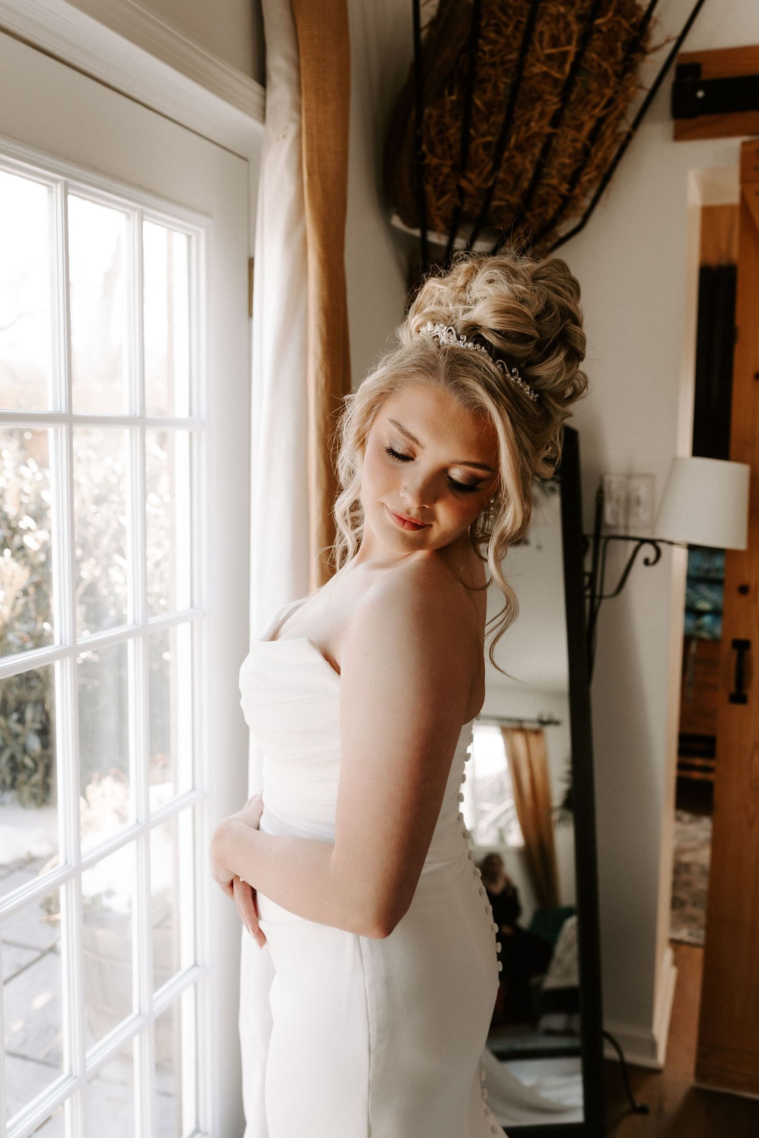 Elegant bride in white gown with updo hairstyle stands by window in soft natural light.