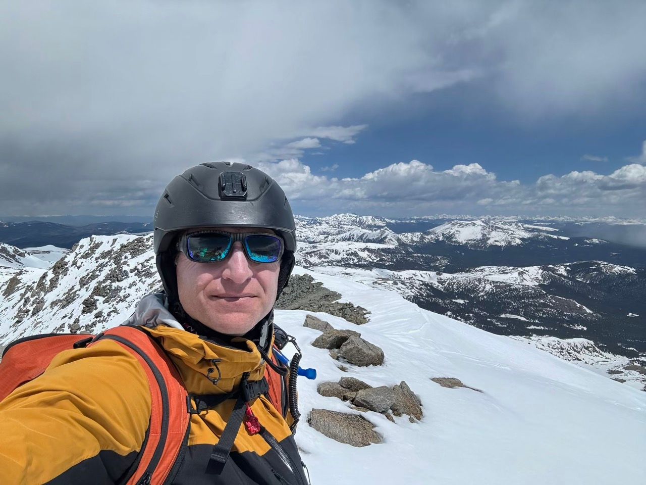 Mountain adventurer takes selfie on snowy summit against dramatic mountain backdrop.