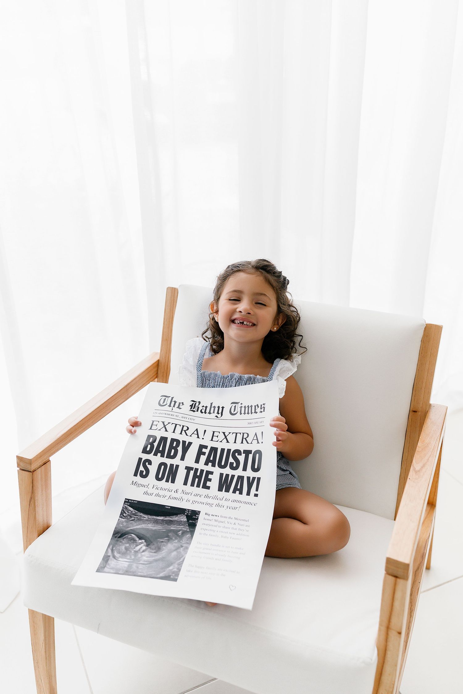 A child sits in a white chair holding a newspaper with a pregnancy announcement featuring an ultrasound photo.