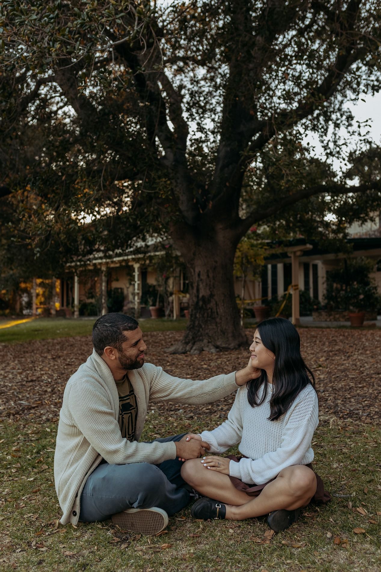 A couple sits together on fallen autumn leaves beneath a large oak tree at the Will Rogers State Park visitor's center.