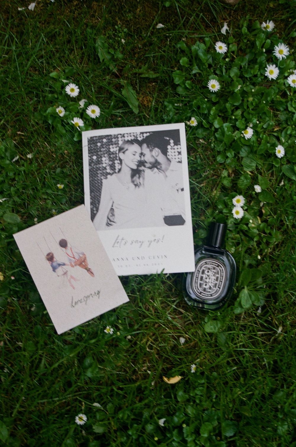 Wedding invitation cards and Diptyque perfume bottle resting on green grass with white daisies.
