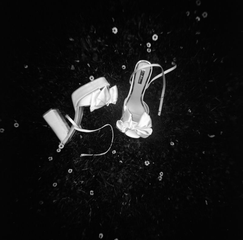 Black and white photo of elegant white high-heel sandals with bow detail, suspended mid-air with water droplets splashing around them.