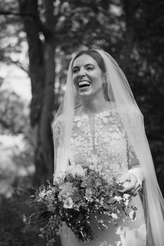 Black and white photo of a laughing bride in lace gown and veil holding a bouquet outdoors.