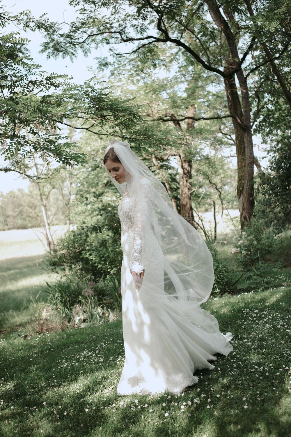 Bride in lace long-sleeve wedding gown and flowing veil standing in lush green garden setting.