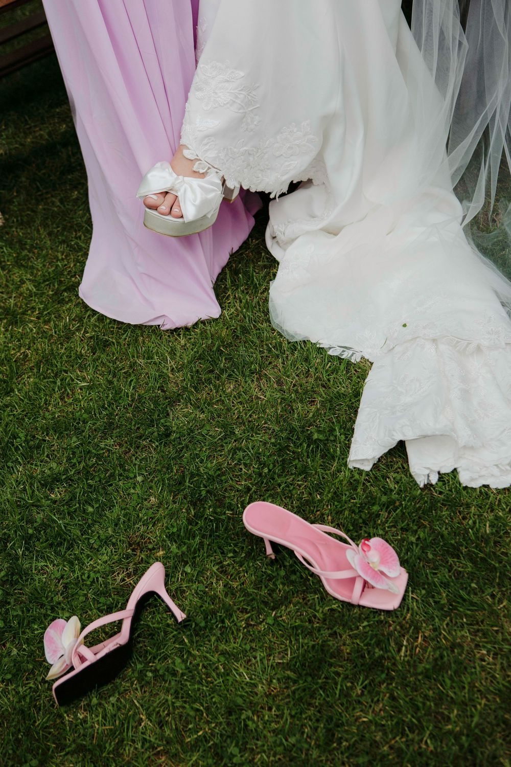 Pink bridal heels and flip flops on grass beside bride in white gown and bridesmaid in lavender dress.