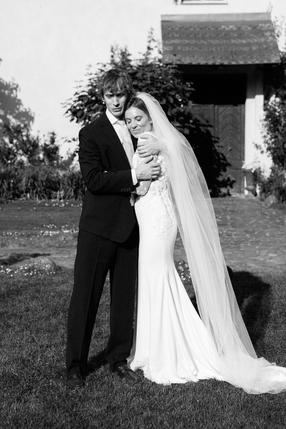 Black and white wedding photo of bride and groom embracing outdoors near a building with lush greenery.