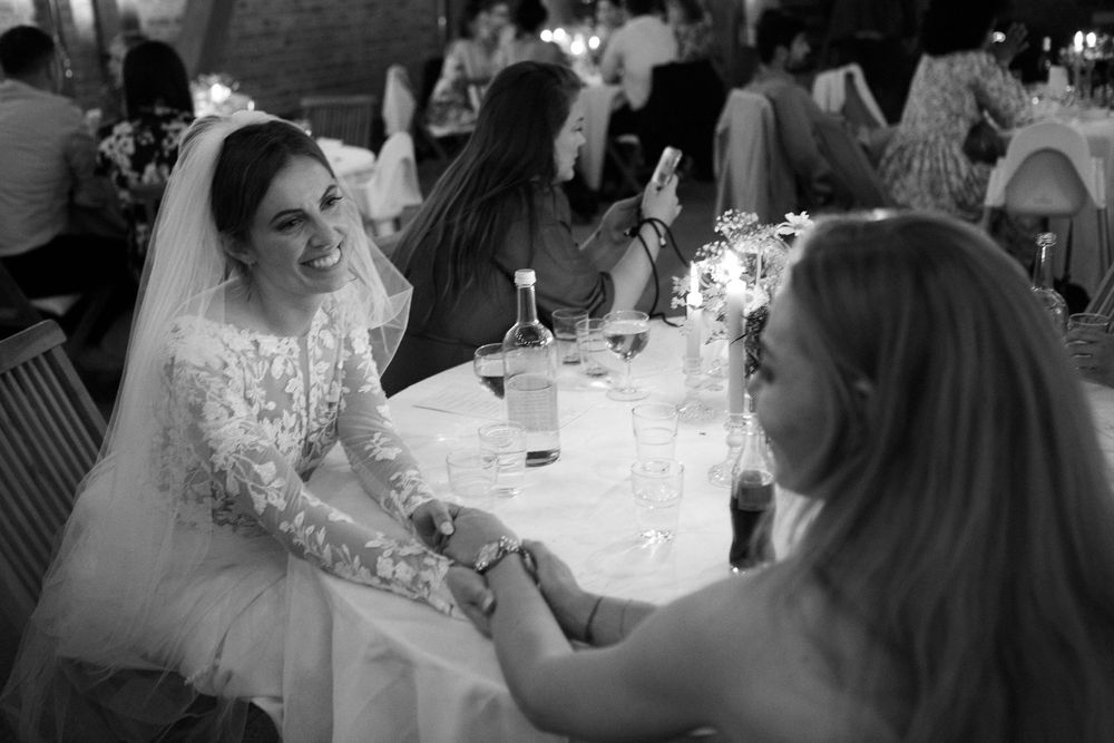 Bride in lace wedding gown smiling and holding hands with guest at reception table in black and white photo.