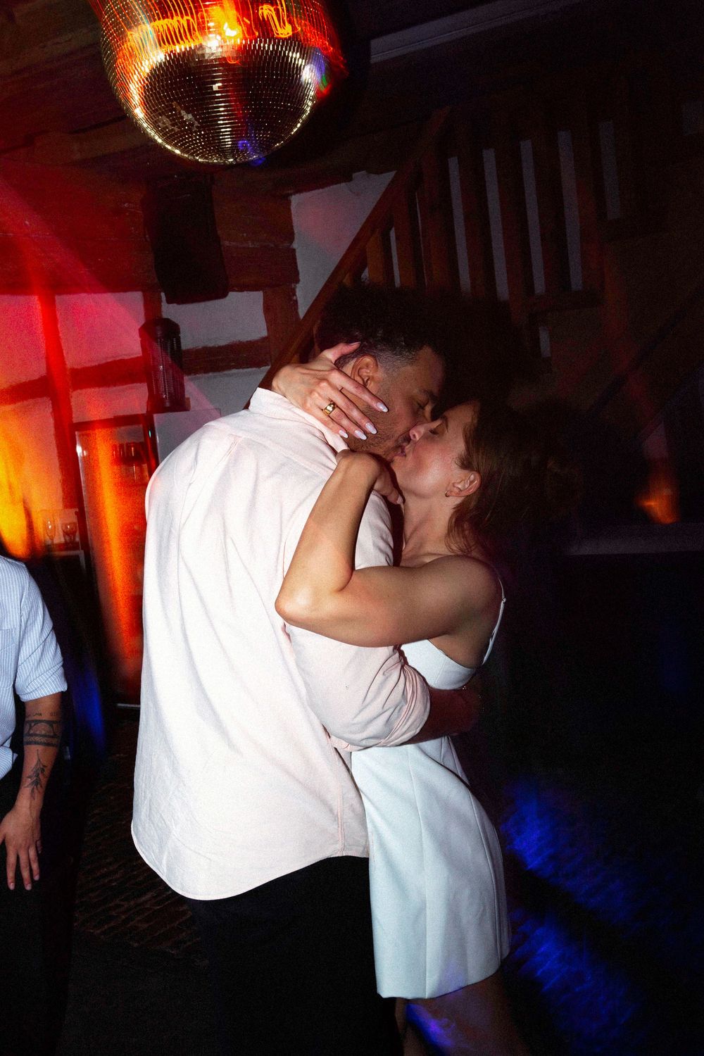 Couple sharing a romantic kiss on a dance floor under a disco ball with colorful lights.
