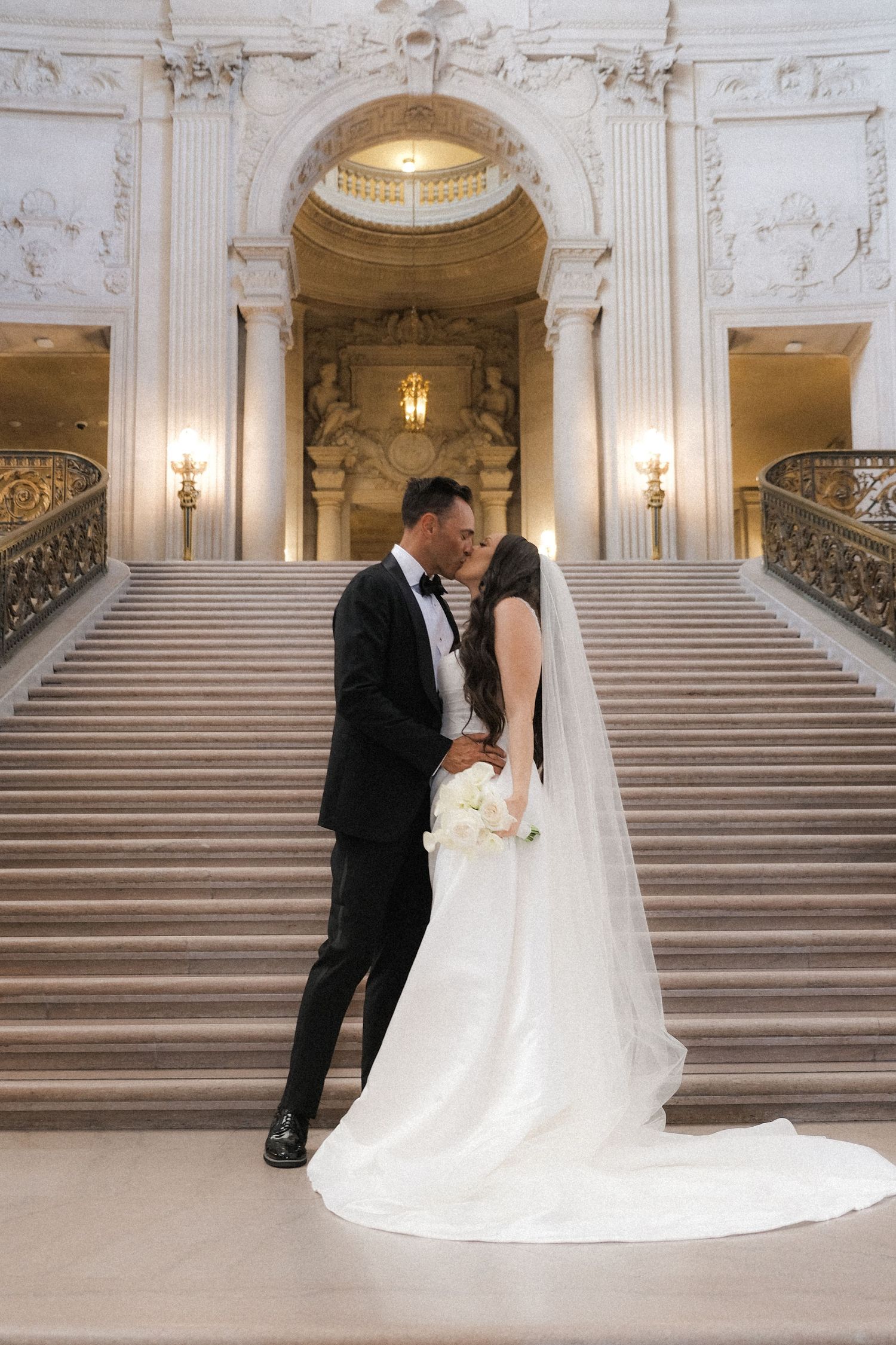 Bride and groom walking hand in hand down ornate grand staircase at San Francisco City Hall wedding.