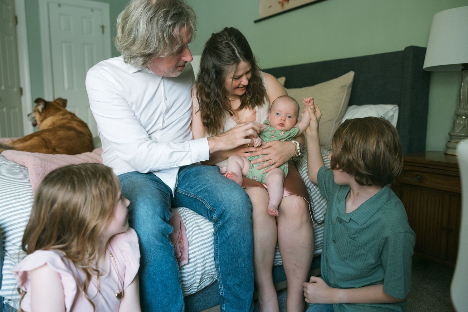 Family gathered around newborn baby on bed, with children and dog in cozy bedroom setting.