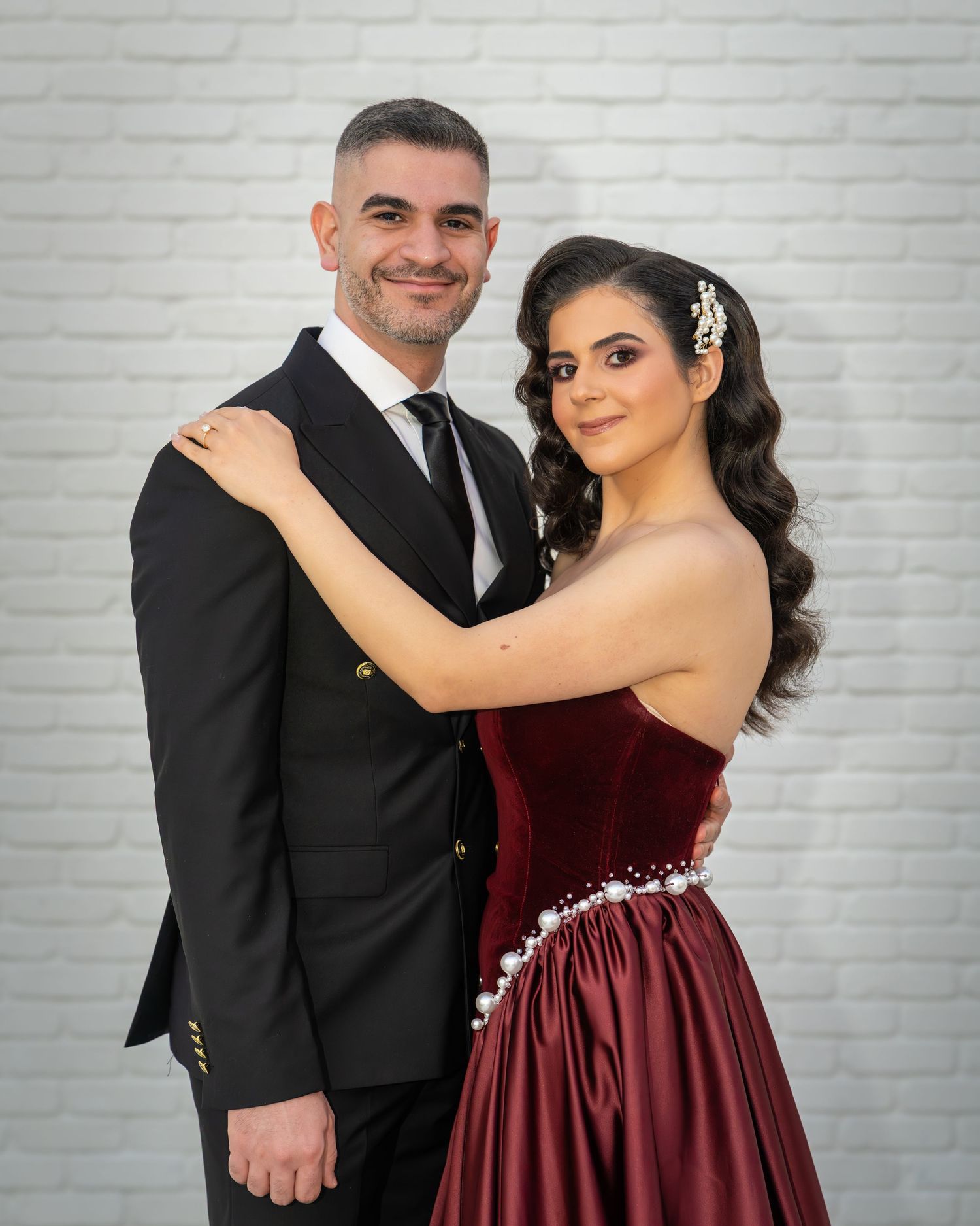 Couple posing elegantly, man in black suit, woman in burgundy velvet strapless gown, against white brick wall.