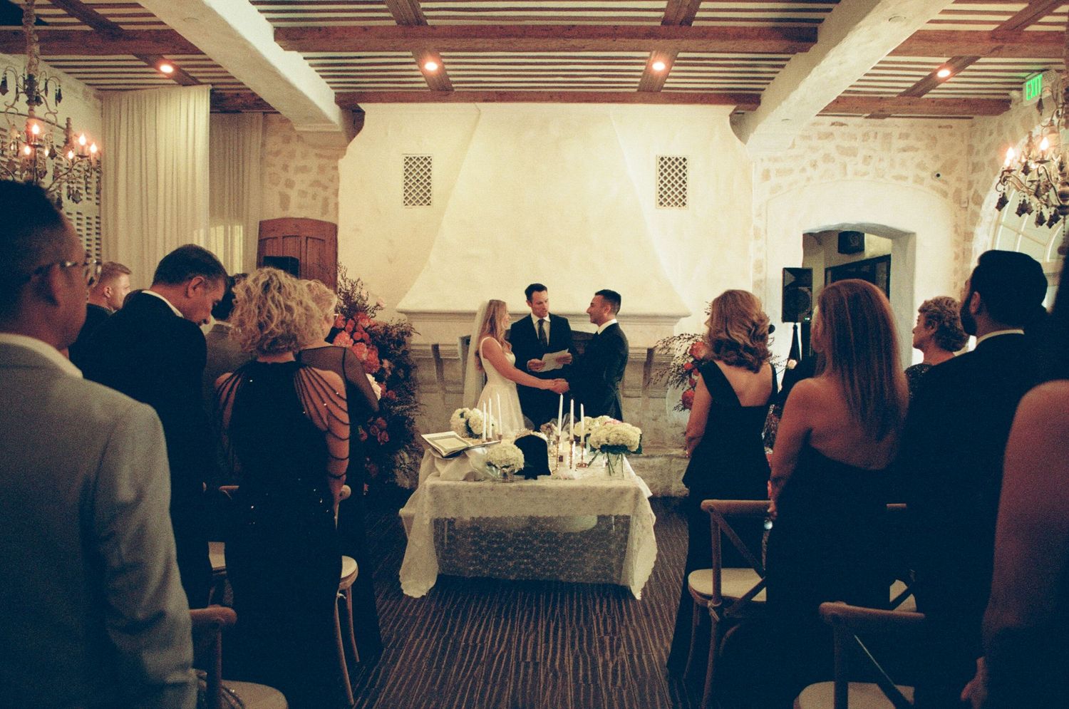 Elegant indoor wedding ceremony with guests standing as couple exchanges vows at decorated altar in chandelier-lit venue.