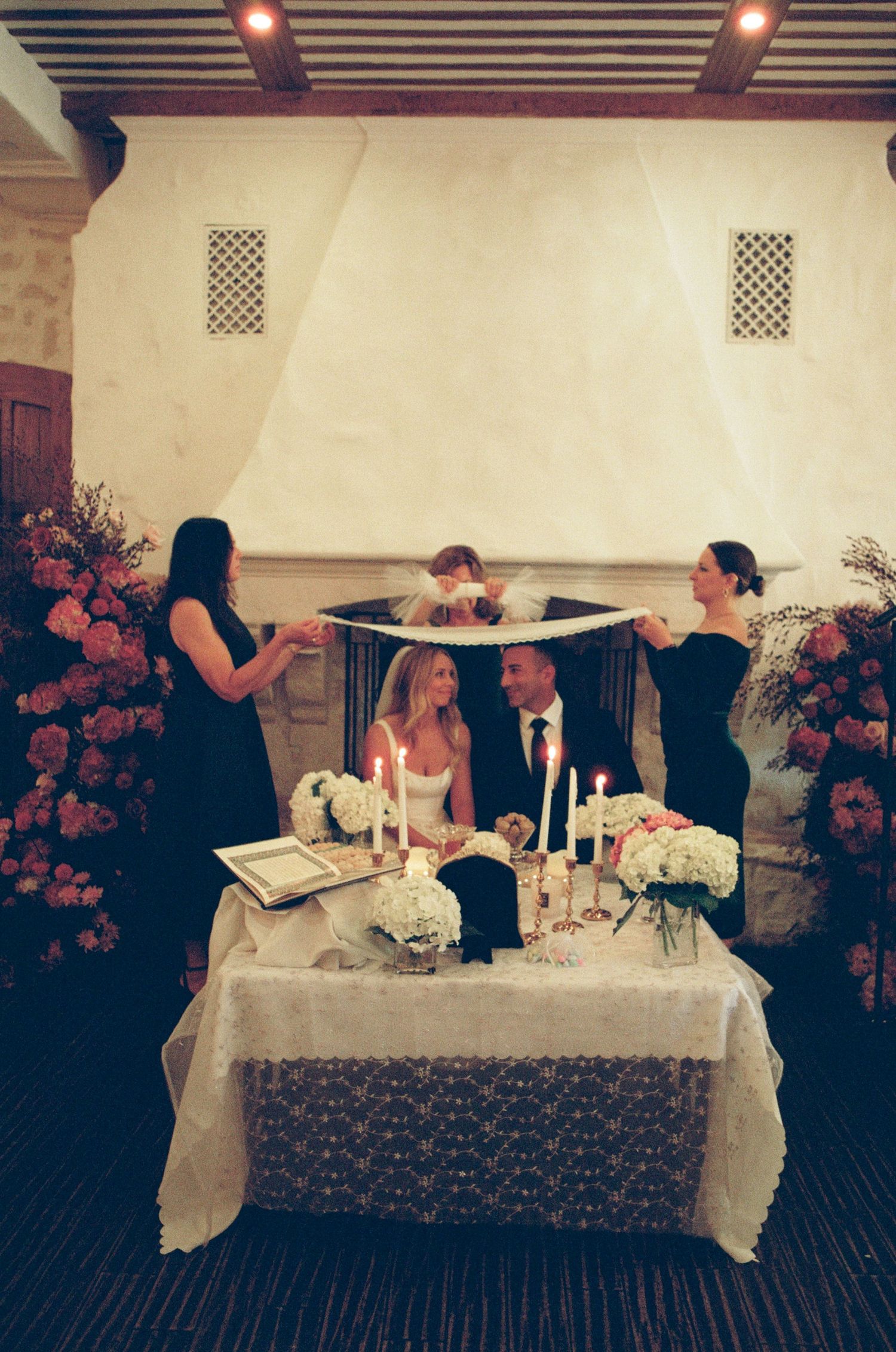 Jewish wedding ceremony under a chuppah with bride and groom at candlelit altar surrounded by pink floral arrangements.