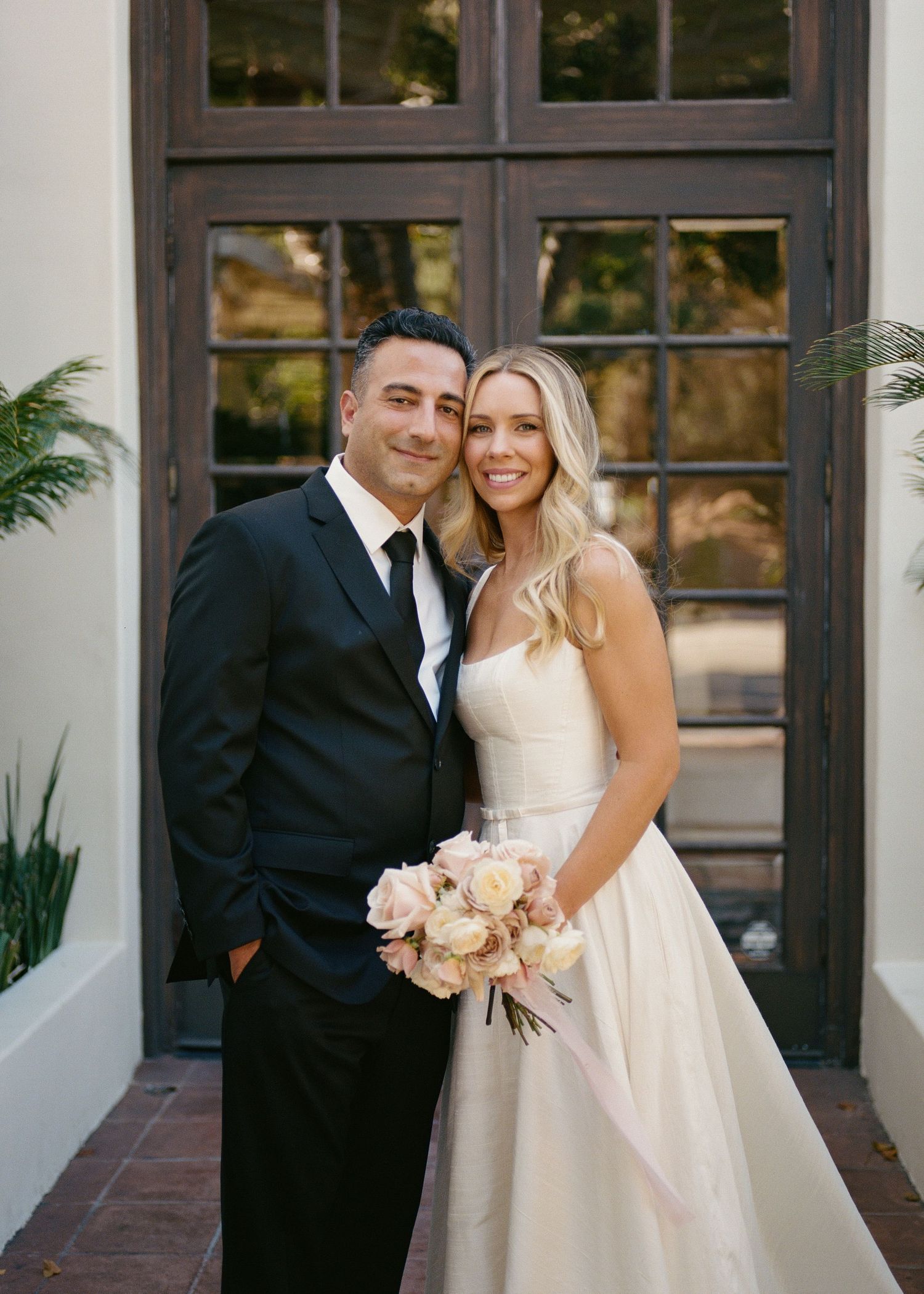 Smiling bride and groom portrait outside wooden doors, bride holding blush pink flower bouquet.