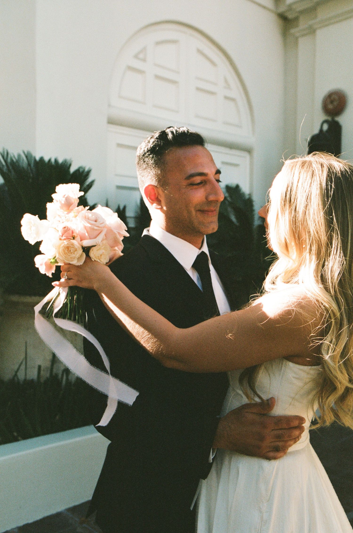 Groom embracing bride holding pink rose bouquet outside white arched church entrance.